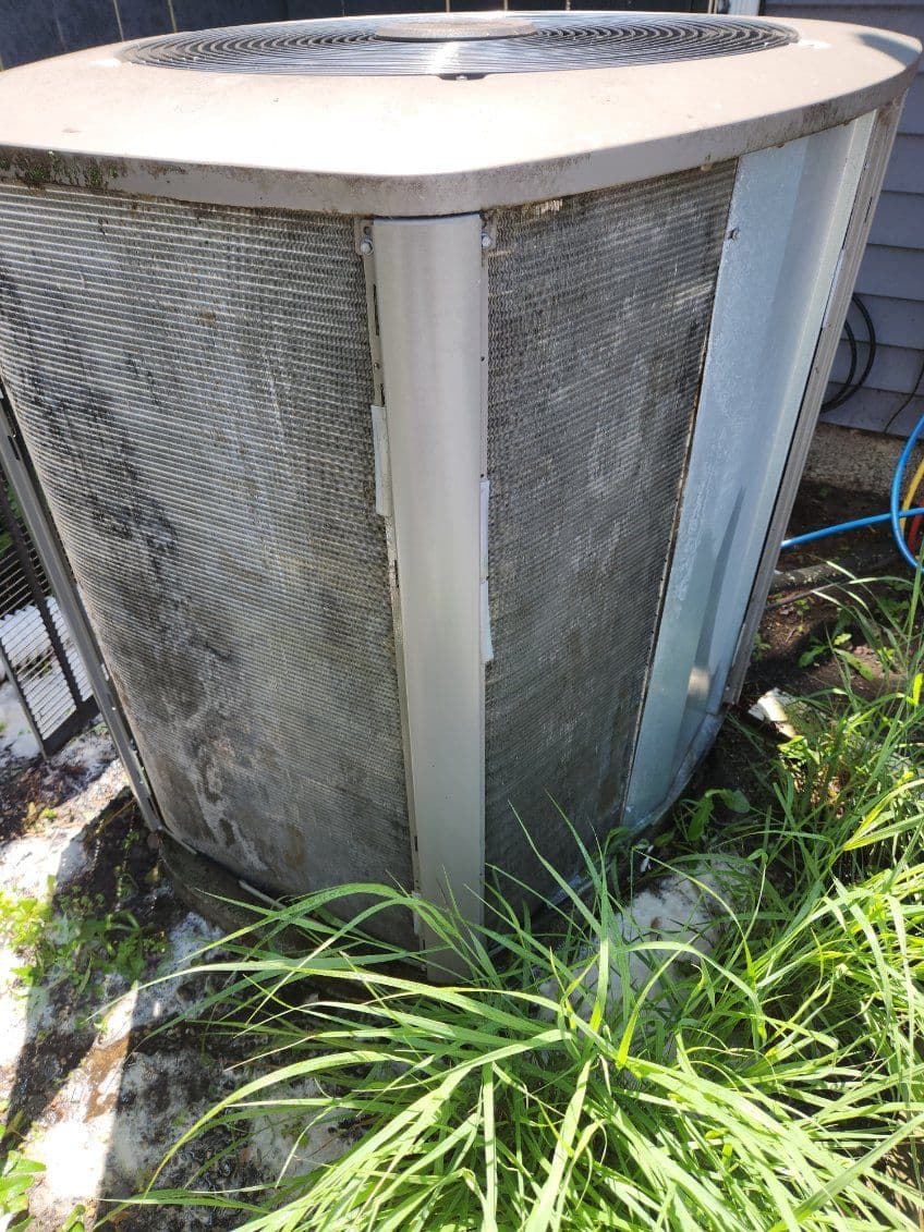 Dirty air conditioning unit surrounded by overgrown grass, indicating maintenance needs.