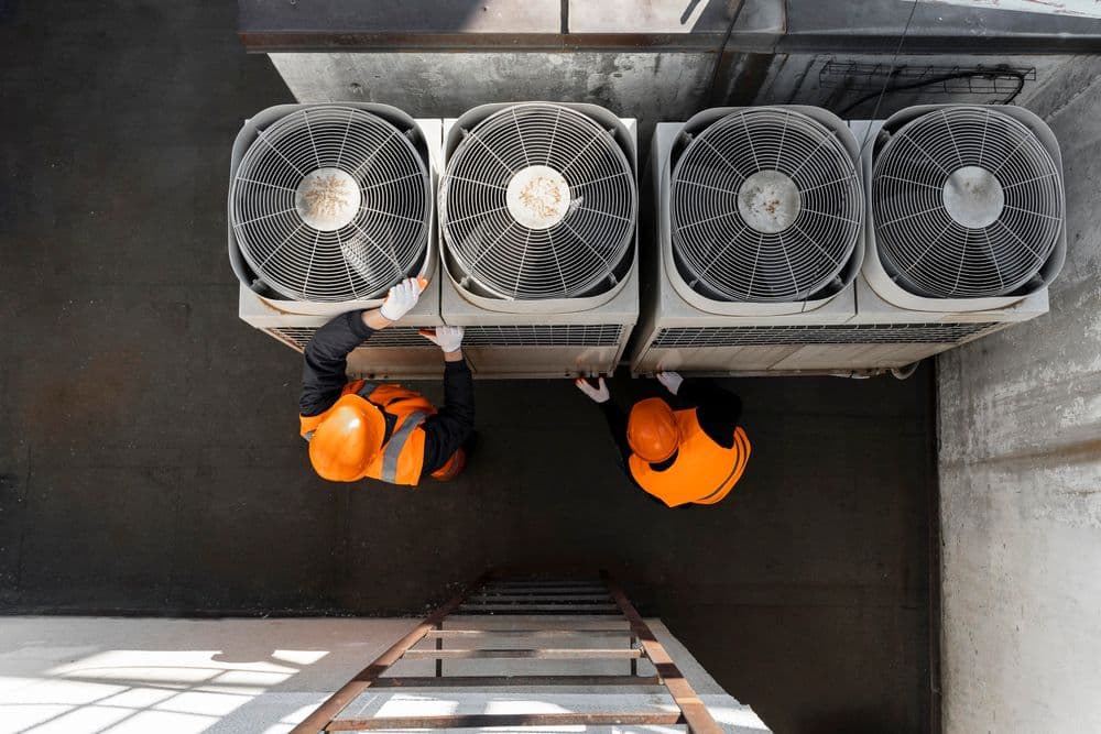 Technicians in safety gear servicing air conditioning units on a rooftop.