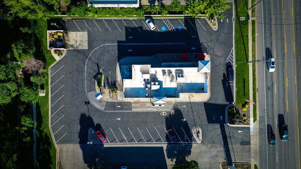 Aerial view of a commercial building with outdoor seating and parking lot. Surrounded by greenery.
