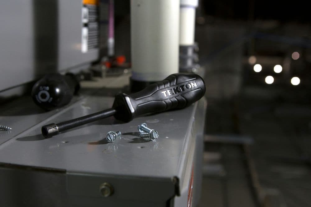 TEKTON screwdriver resting on an electrical panel with screws nearby, showcasing tool detail.