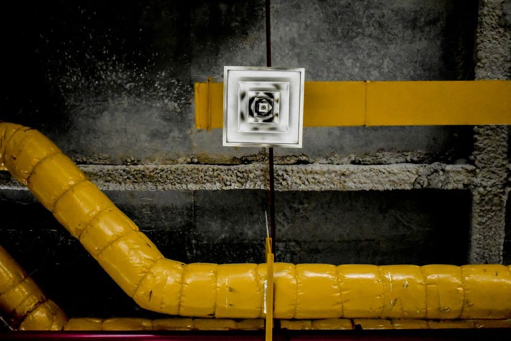 Ceiling ductwork with yellow insulation and a ventilation grille in an industrial setting.