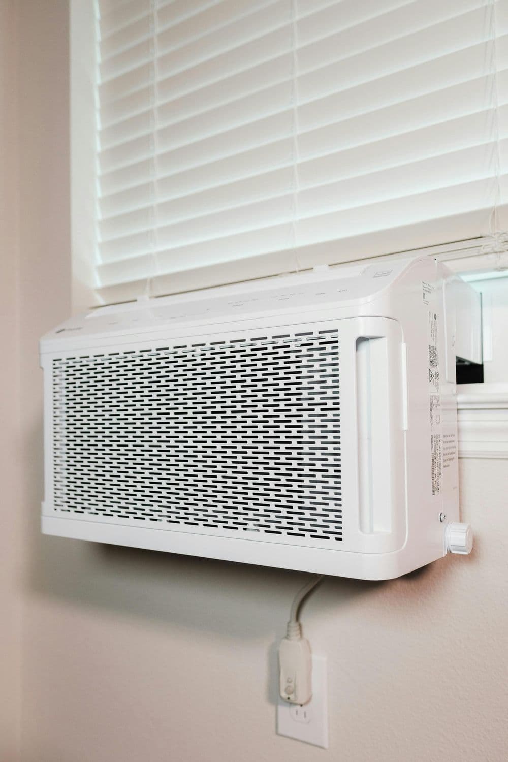 White window air conditioner installed below a window with closed blinds.